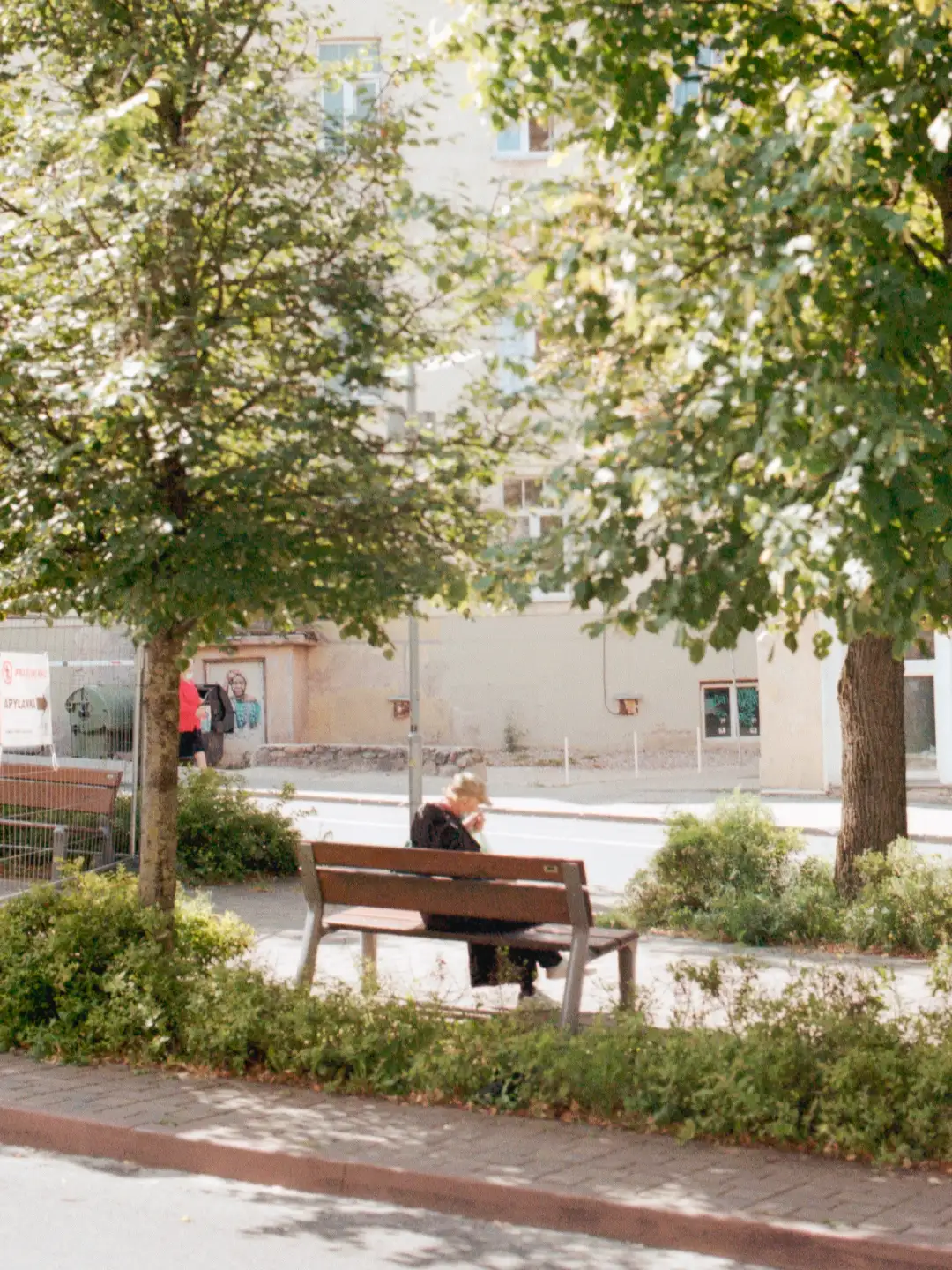 Film style image of a person sitting on a bench in a small park surrounded by trees. Created and photographed by Hiroku