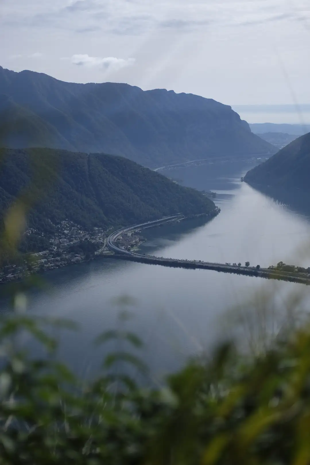 A highview photo of a road leading over a lake inbetween a couple of mountains. Created and photographed by Hiroku